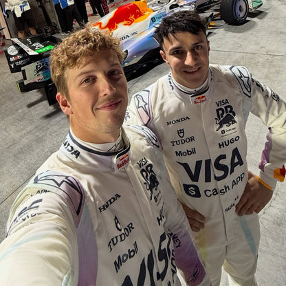 Two Formula 1 drivers in white racing suits with various sponsor logos taking a selfie in front of a Red Bull Racing car.