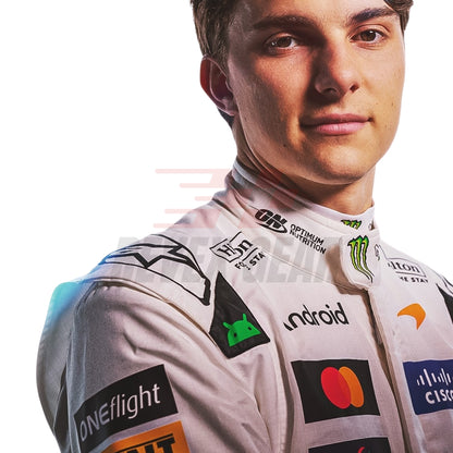 A close-up portrait of a male race driver, Oscar Piastri, wearing a McLaren 2026 British GP white F1 racing suit covered in sponsor logos, including Android, Mastercard, Optimum Motion, and Monster Energy, against a plain white background.